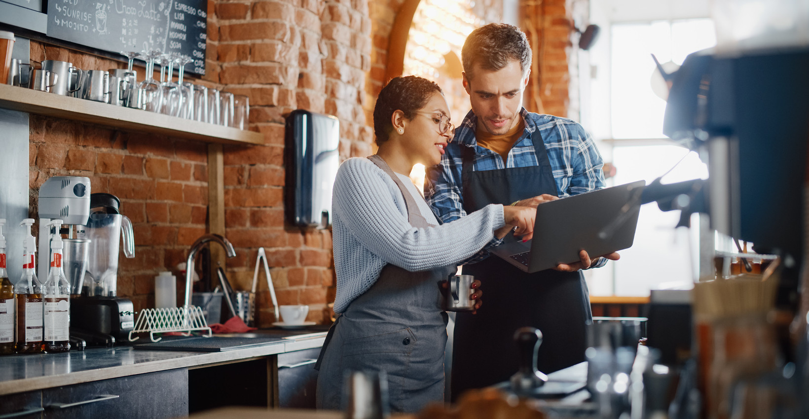 A man an dwoman looking at a laptop behind the counter of a coffee shop
