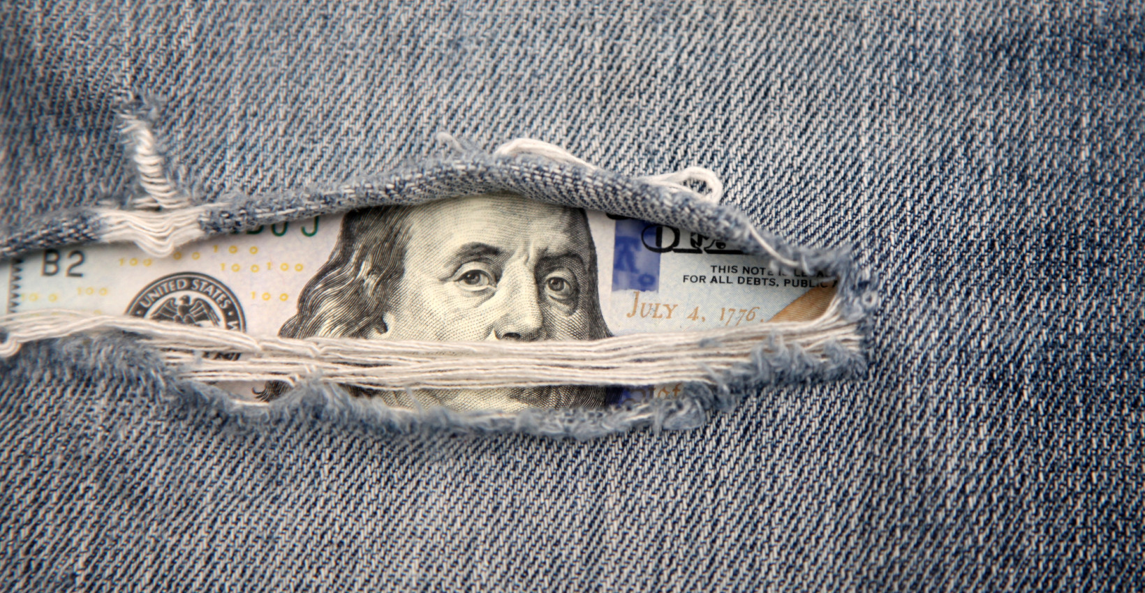 a hundred dollar bill peaks out of ripped jeans