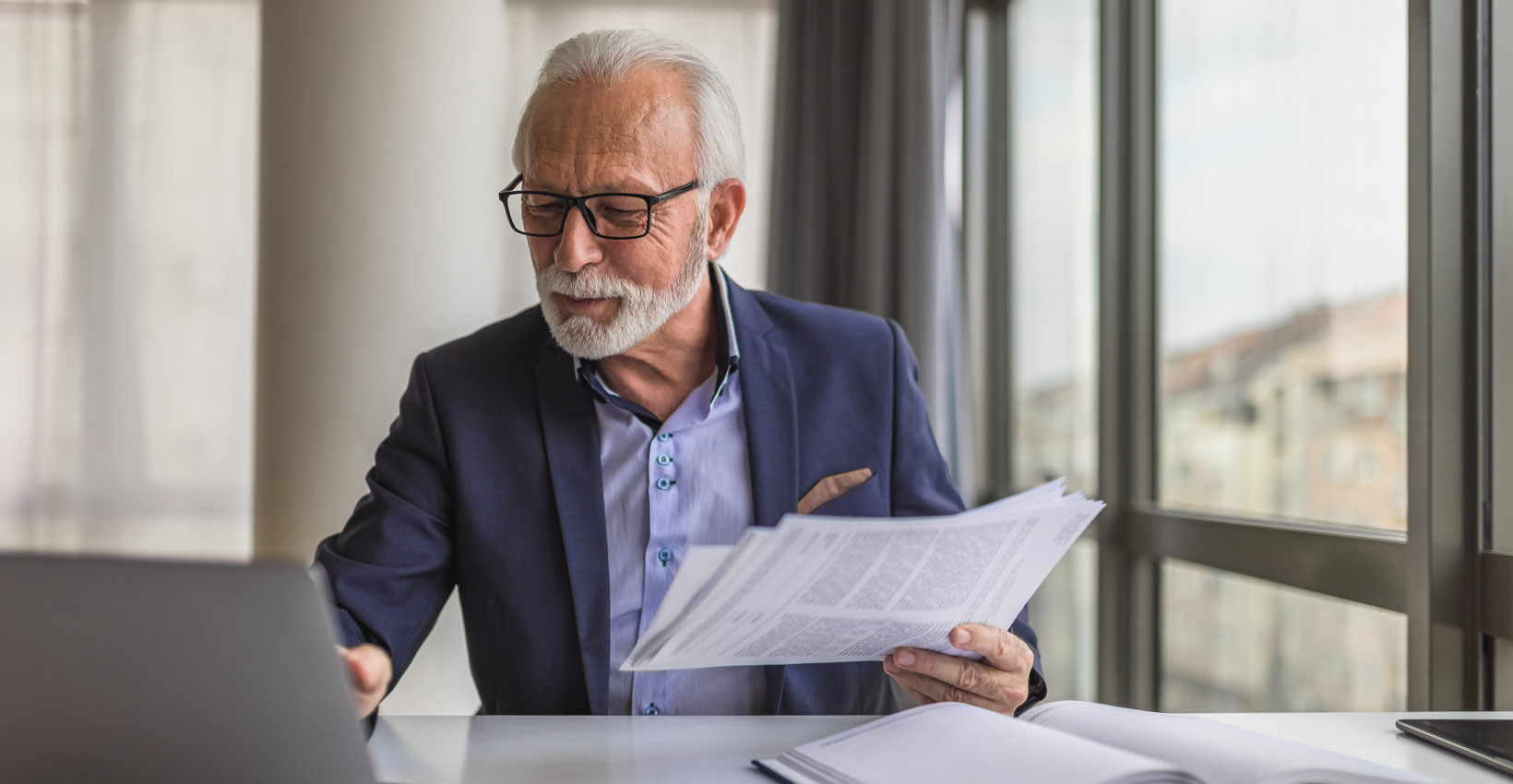 an older man wearing a blue suit sits at a computer with papers in his hands