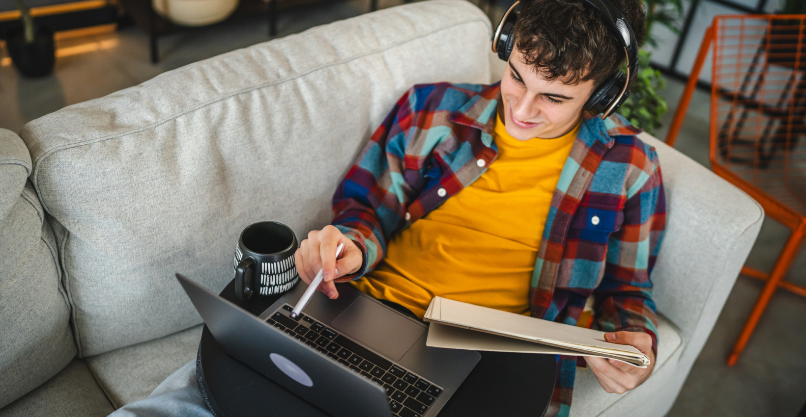a teen lays on the couch with his laptop on his lap and holds a notebook while wearing headphones