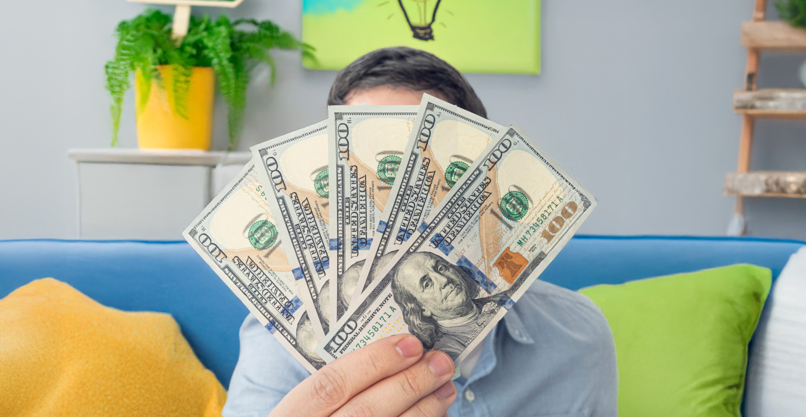 a person wearing a blue shirt holds up five 100 dollar bills while sitting on a couch