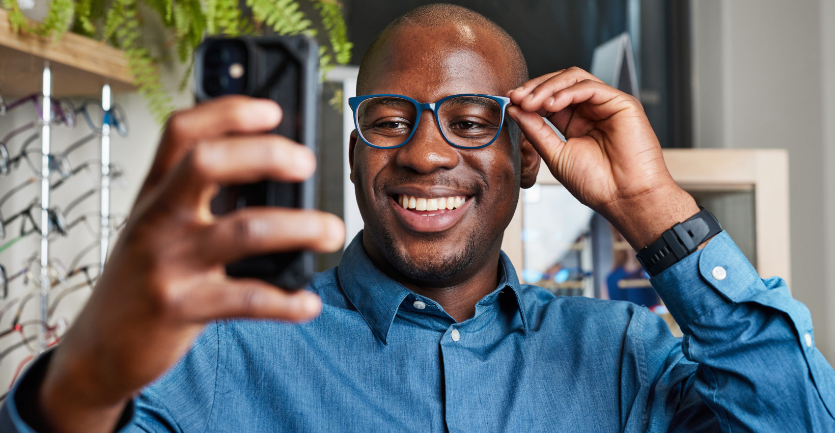 a black man wearing a blue button up shirt smiles while taking a selfie on his phone
