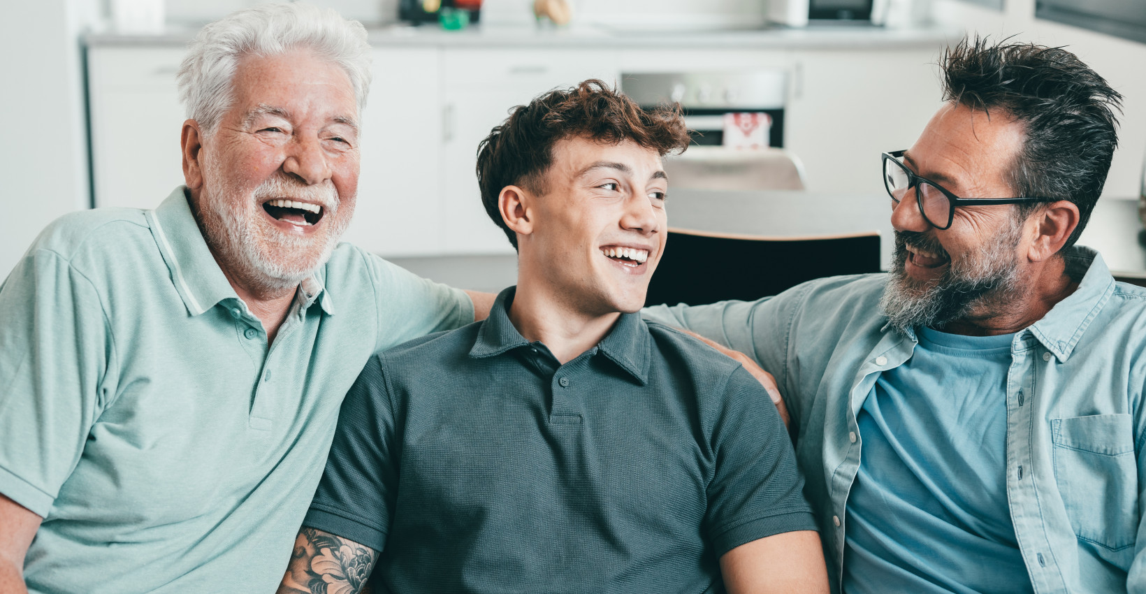 a young, middle-aged, and older man sit together on a couch laughing