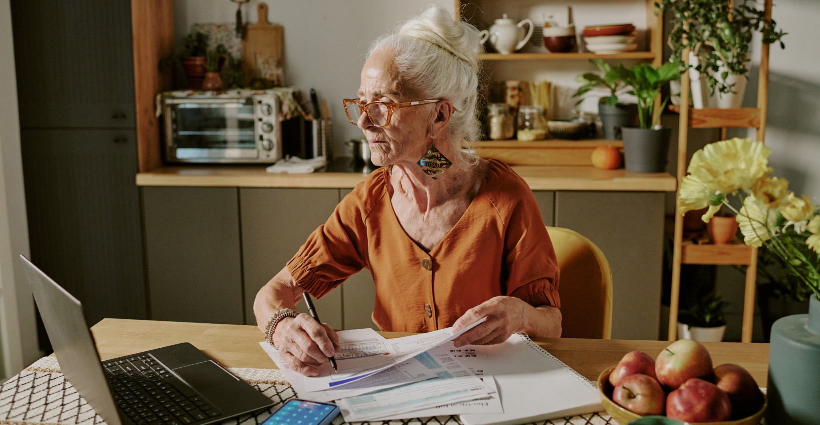 an elderly woman sits at a desk with paperwork and a laptop
