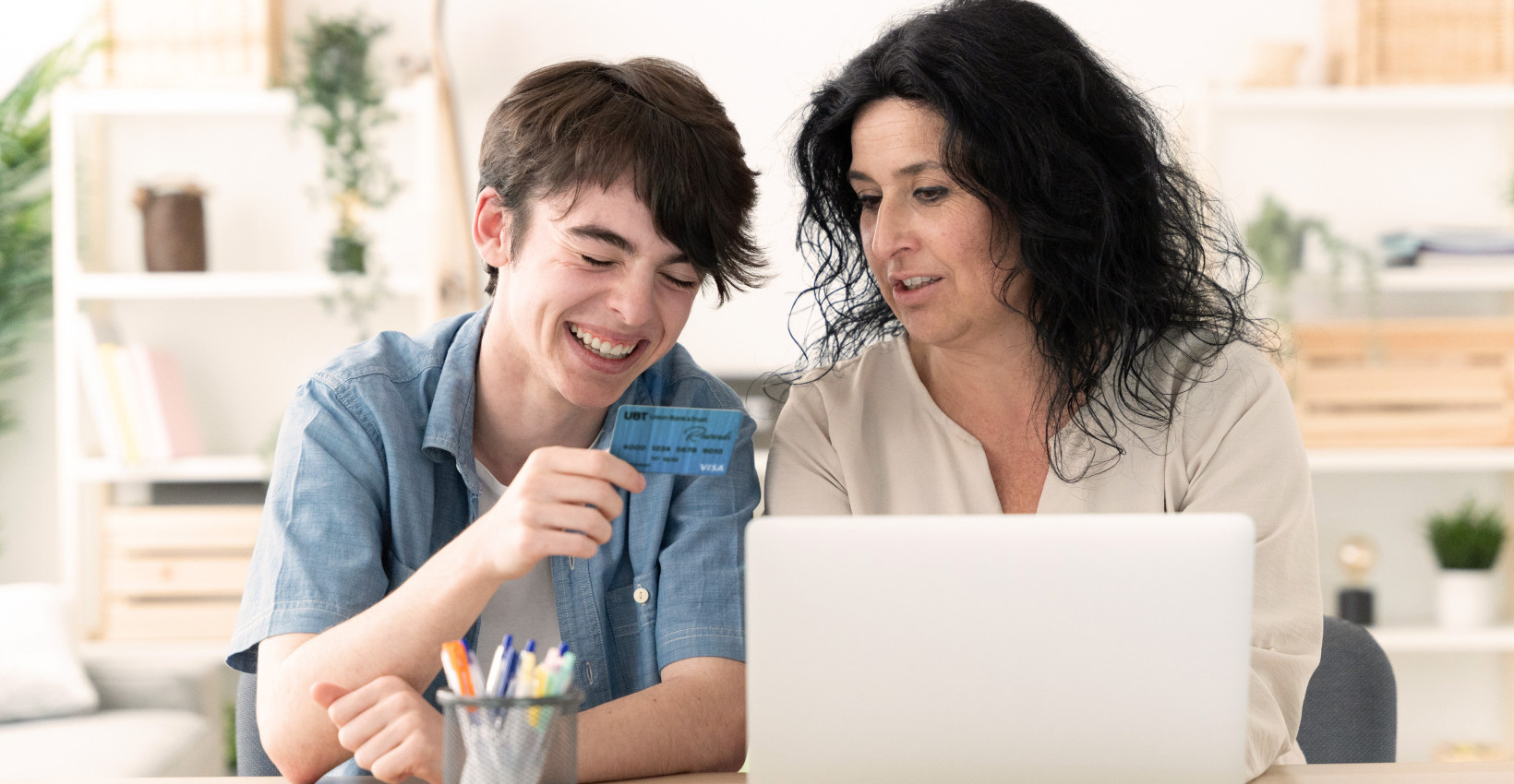 a mother and son sit at a laptop together laughing. the son holds a credit card