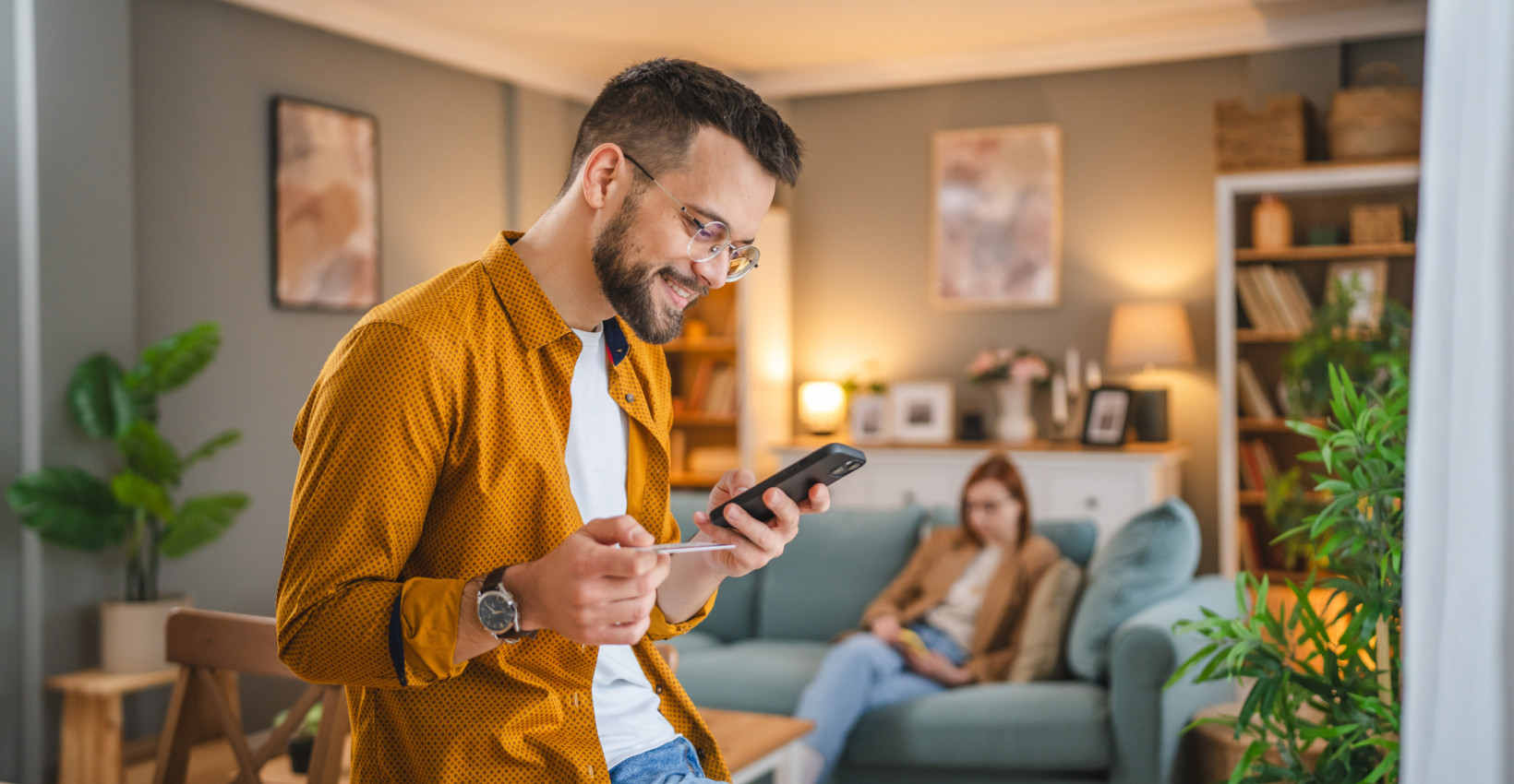 a man leans on a table holding his phone and debit card while a woman sits on the couch behind him