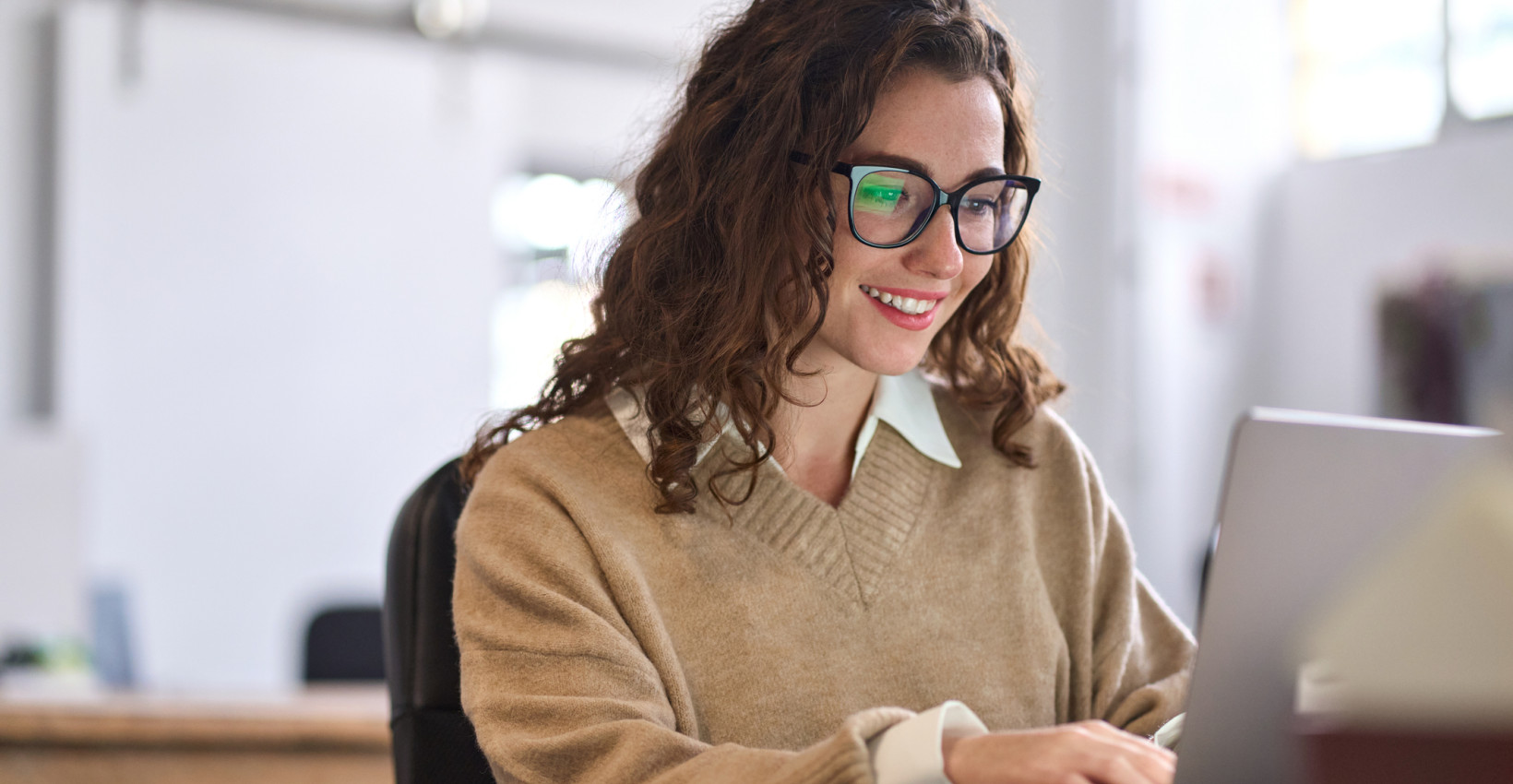 a girl with curly hair and glasses sits at a laptop smiling