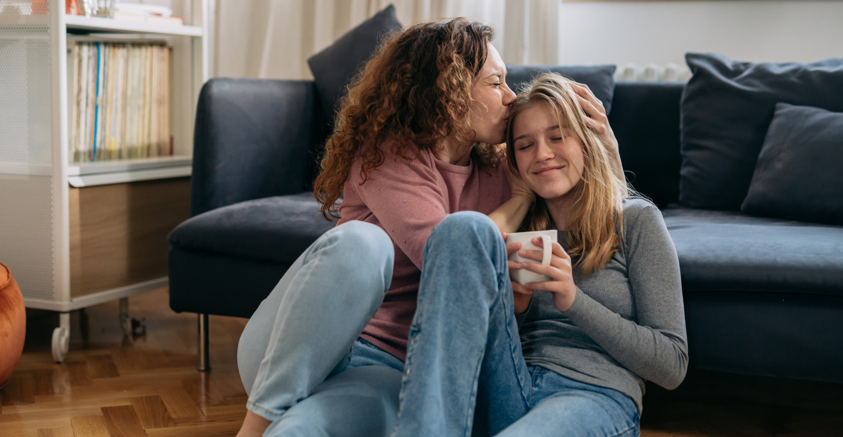 a mom and daughter sit together on the floor leaning up on the couch. the mom kisses the daughters head.