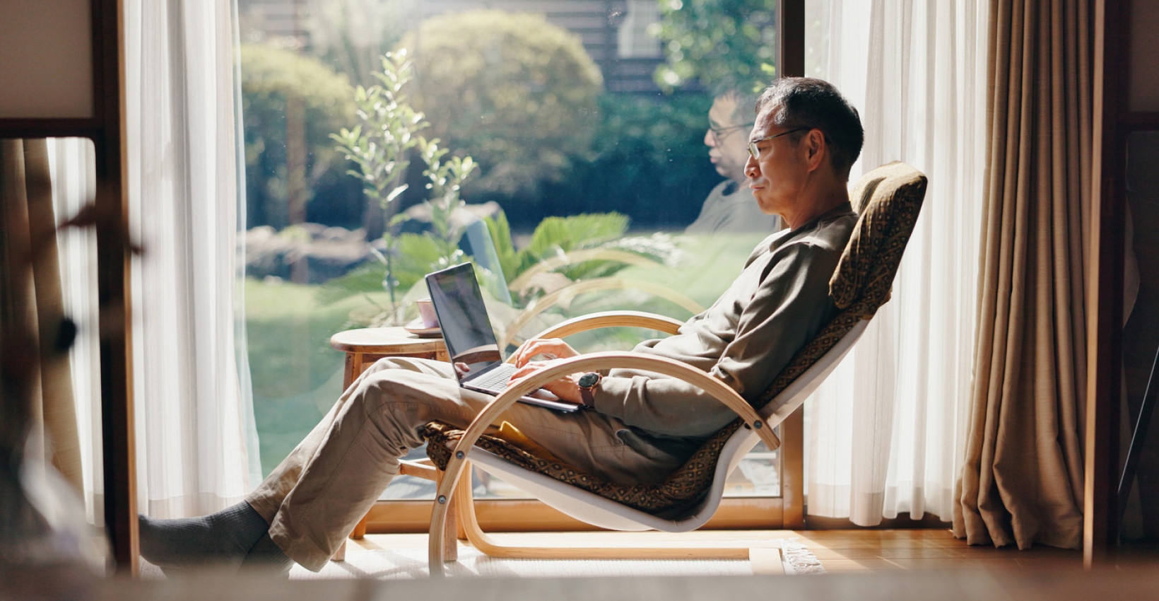 a man sits in a rocking chair by a window with his laptop