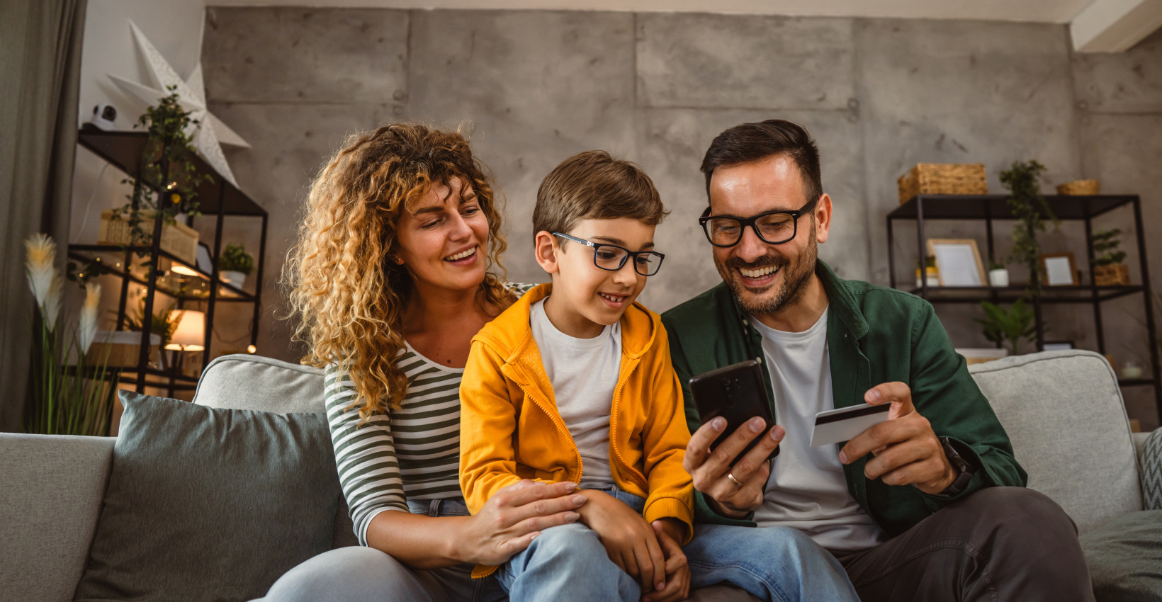 a mom and dad sit with their son on a couch looking at a phone and debit card