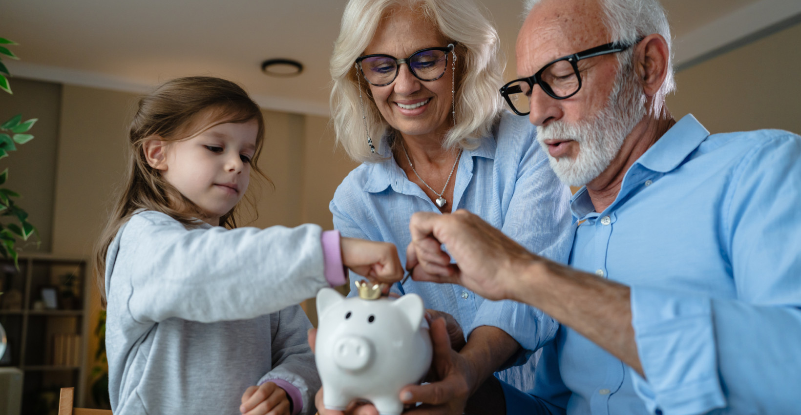 a grandma and grandpa help their granddaughter put coins into a white piggy bank