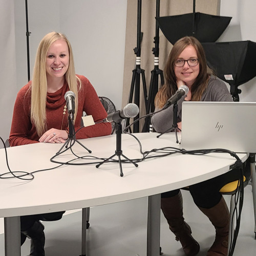 Two women sitting in a podcast studio smiling at the camera