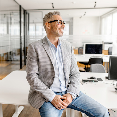 a guy wearing a blazer and jeans sits on a table smiling looking away from camera