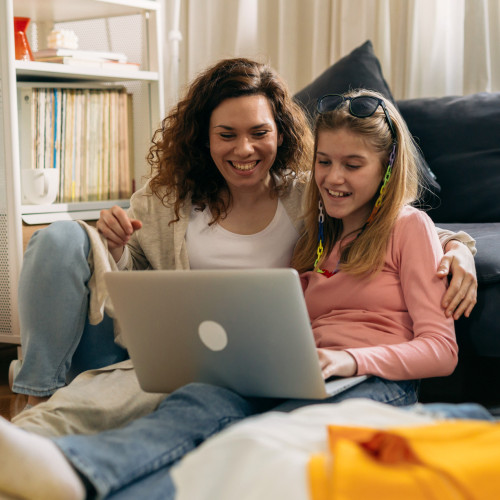 a mom and daughter sit on the couch together looking at a laptop