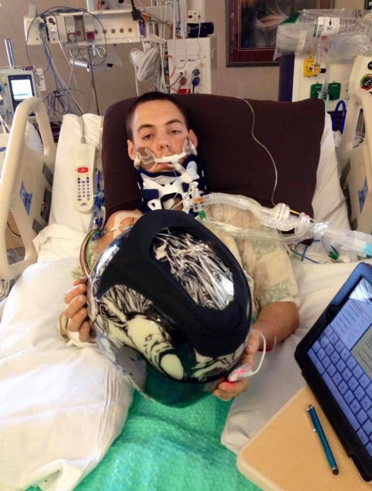 A man in serious condition on a hospital bed holding up the motorcycle helmet that saved his life