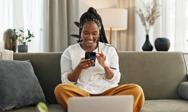 A woman sits on a her couch with her laptop in front of her while holding her phone