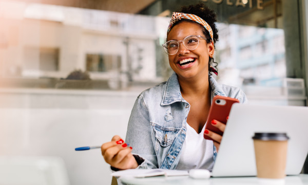 a young black woman smiles while sitting at a table with a laptop and coffee
