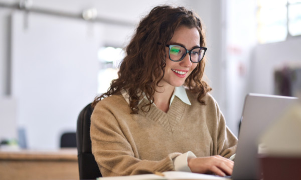 a girl with curly hair and glasses sits at a laptop smiling