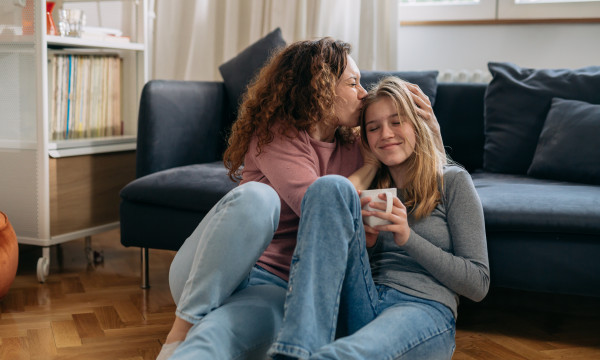 a mom and daughter sit together on the floor leaning up on the couch. the mom kisses the daughters head.
