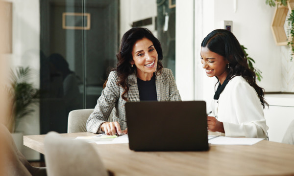 two women sit behind a laptop together smiling